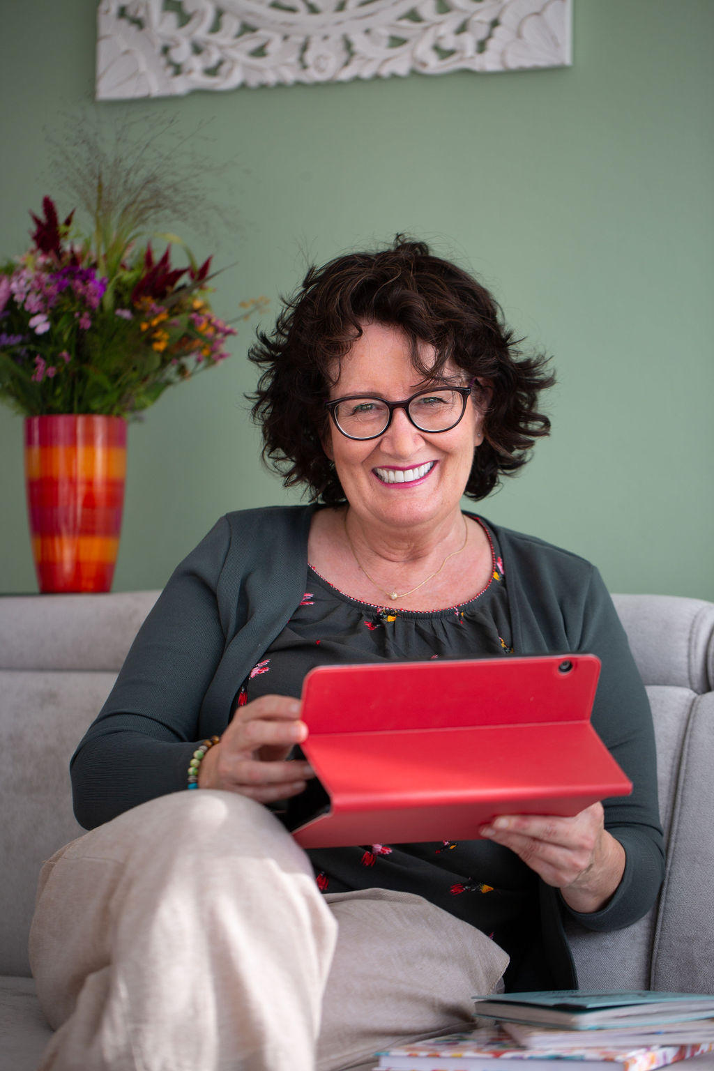 Regina mit Lachen im Gesicht sitzt vor einem Blumenstrauß und hat ein Tablet mit rotem Cover in der Hand